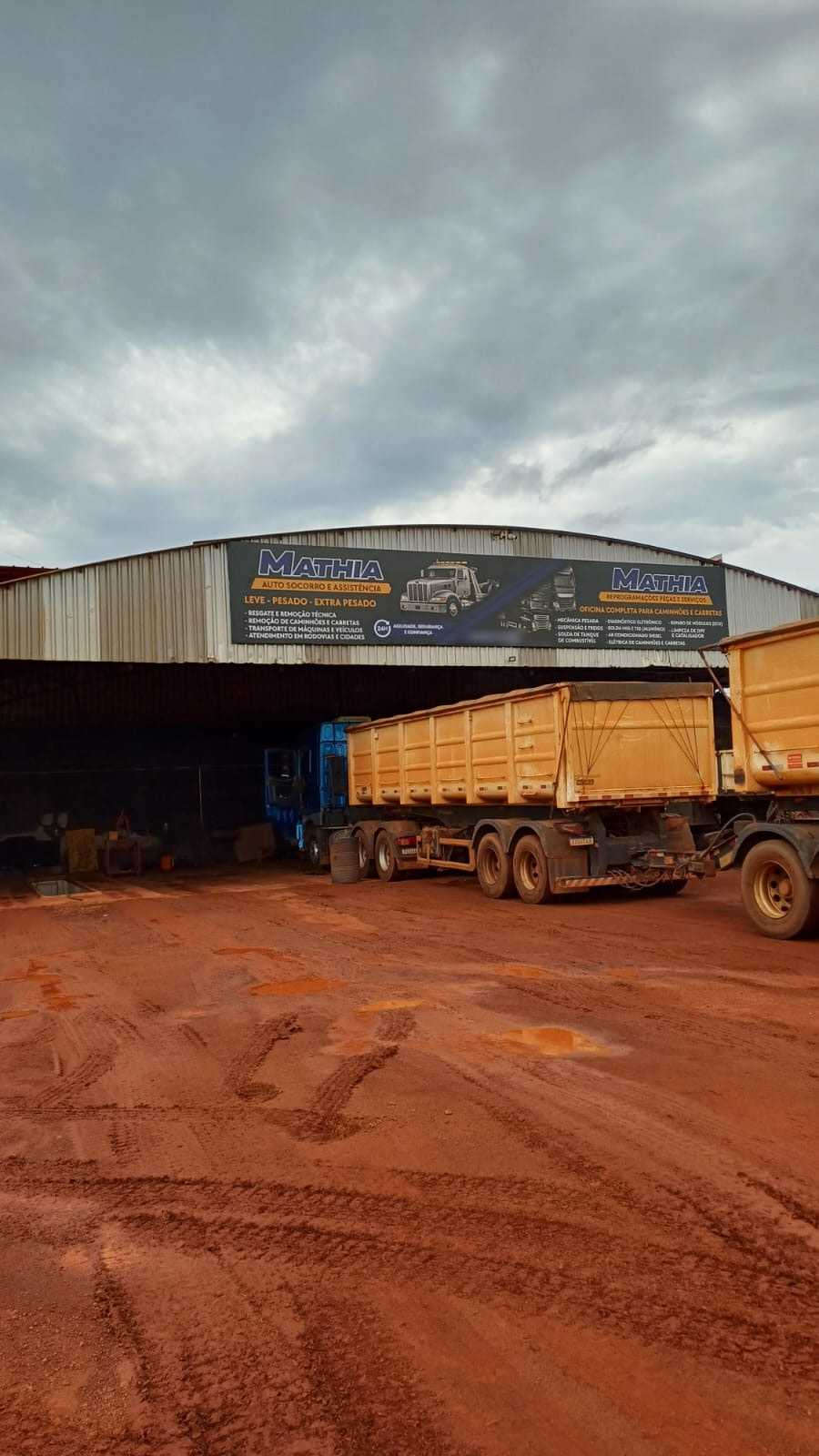 Galpão da Mathia com as duas placas novas e uma carreta basculante amarela saindo da oficina, sob céu nublado (21/10/2026)